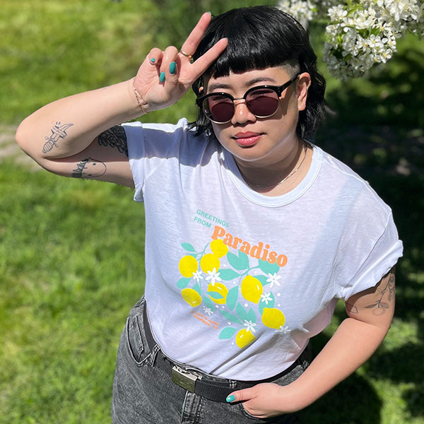 Person wearing the Paradiso T-Shirt with bright lemon print, sunglasses, and teal nail polish outdoors in sunny setting