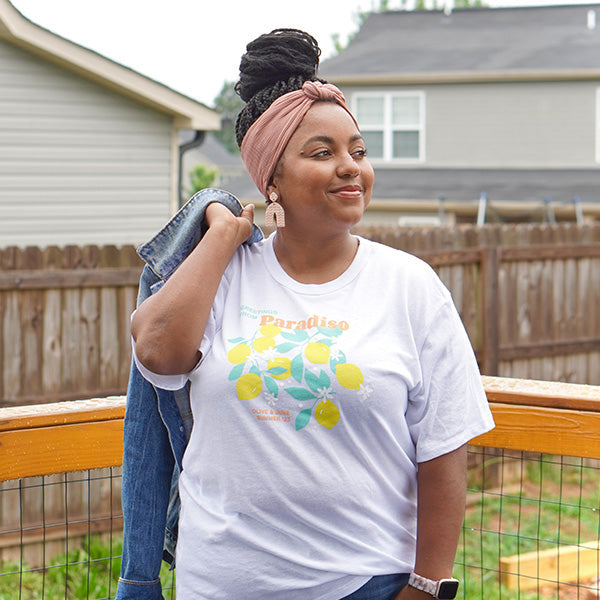 Smiling woman outdoors wearing the Paradiso T-Shirt with lemon and leaf design holding a denim jacket on a wooden deck