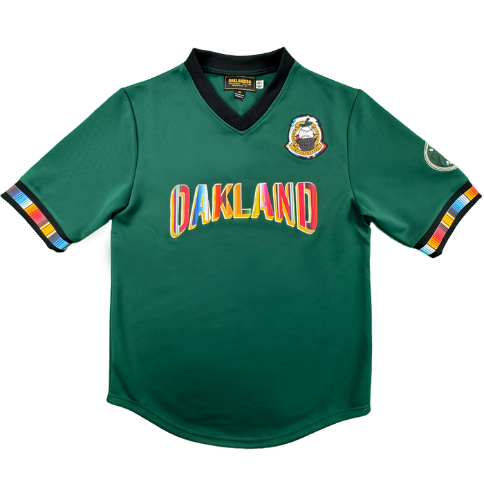 Introducing the Peloteros Jersey by Oakland Ballers: a vibrant green sports jersey inspired by the Oakland Ballers baseball team, featuring "OAKLAND" in multicolored letters across the front, a colorful badge on the left chest, an oval patch on the right sleeve, and black-trimmed sleeves and collar accented with multicolored stripes—a lively tribute to Pioneer League style.