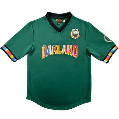 Introducing the Peloteros Jersey by Oakland Ballers: a vibrant green sports jersey inspired by the Oakland Ballers baseball team, featuring "OAKLAND" in multicolored letters across the front, a colorful badge on the left chest, an oval patch on the right sleeve, and black-trimmed sleeves and collar accented with multicolored stripes—a lively tribute to Pioneer League style.