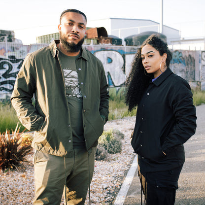 A man and a woman stand outdoors, posing confidently in front of a graffiti-laden wall. The man wears a green water-resistant jacket in men's size and khaki pants, his hair neatly braided. The woman, with long curly hair, is dressed in a black Premium Coaches Jacket by Oaklandish and matching pants, accessorized with large hoop earrings. They both have serious expressions on their faces.