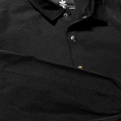 Close-up of the Oaklandish Premium Coaches Jacket in black. The image showcases the collar and part of the upper section, focusing on the metallic buttons. The matte fabric appears smooth. The top button is fastened, while the second button remains unbuttoned. The sleeve is partially visible, hinting at its men's sizing.