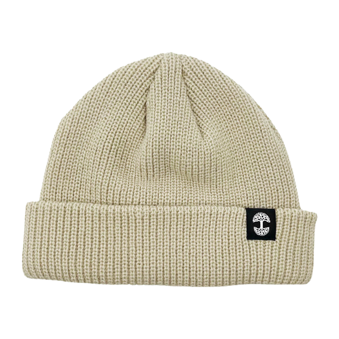 The Ramos Beanie by Oaklandish is depicted, showcasing headwear at its finest with its beige knit and folded brim. The brim features a distinctive black rectangular patch adorned with a white circular cross logo. Made from woven acrylic, the textured material likely provides warmth and comfort. The beanie boasts a simple, classic design that exudes timeless style.