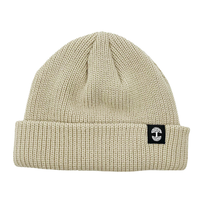 The Ramos Beanie by Oaklandish is depicted, showcasing headwear at its finest with its beige knit and folded brim. The brim features a distinctive black rectangular patch adorned with a white circular cross logo. Made from woven acrylic, the textured material likely provides warmth and comfort. The beanie boasts a simple, classic design that exudes timeless style.