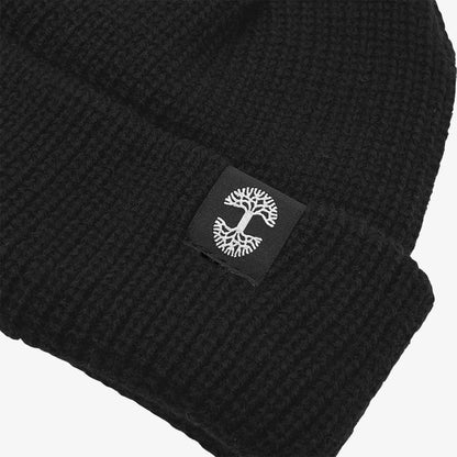 Here's a close-up view of the Oaklandish Ramos Beanie folded at the brim. This black knit headwear showcases a small black square patch with an embroidered white tree design on the fold. The texture of its woven acrylic fabric is clearly visible, emphasizing its ribbed pattern.