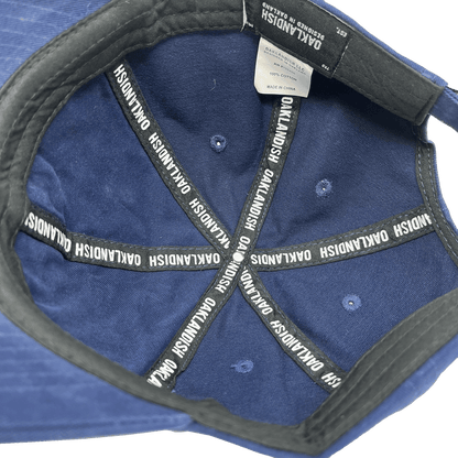 The interior view of the Regal Crest Dad Hat in dark blue from Oaklandish shows eight black straps evenly radiating from the center, each imprinted with the white Oaklandish logo. The black inner band and a white tag are visible, indicating that the hat is made in China from 100% cotton and includes an adjustable strap for a perfect fit.