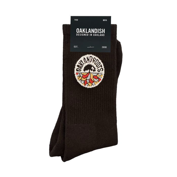 The Roots SC Socks by Oakland Roots SC are black cotton crew socks with a colorful tree and roots embroidered patch. The label reads "OAKLANDISH Designed in Oakland," “one size fits most,” and includes "EST. 2000" on the packaging.