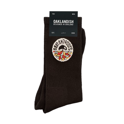 The Roots SC Socks by Oakland Roots SC are black cotton crew socks with a colorful tree and roots embroidered patch. The label reads "OAKLANDISH Designed in Oakland," “one size fits most,” and includes "EST. 2000" on the packaging.
