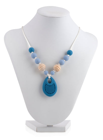 The Nuby Silicone Teething Necklace features blue, beige, and white soft silicone round beads with a large blue teardrop pendant, displayed on a white jewelry stand.