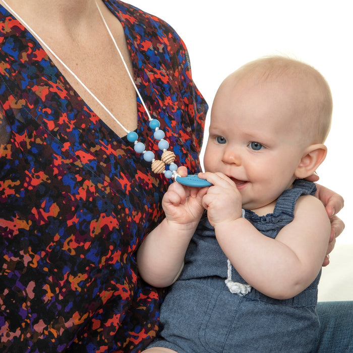 A baby in a blue romper sits on an adult's lap, happily chewing on the Nuby Silicone Teething Necklace worn by the adult, who is dressed in a playful multicolored top ideal for soothing teething babies.