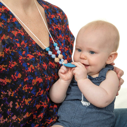 A baby in a blue romper sits on an adult's lap, happily chewing on the Nuby Silicone Teething Necklace worn by the adult, who is dressed in a playful multicolored top ideal for soothing teething babies.