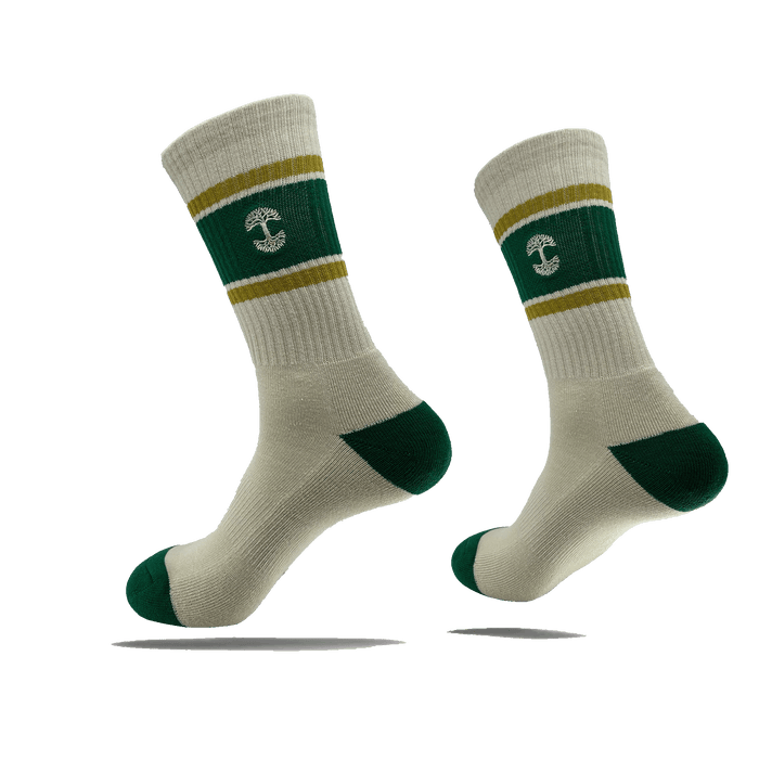 Introducing the Striped Classic Sock by Oaklandish: a pair of beige crew socks featuring green heels, toes, and cuffs. The socks sport a green band around the lower leg area, accented with two thin yellow stripes above and below it. At the center of this green band is a white anchor symbol. Crafted from 70% cotton, these stylish socks are available in adult sizes. They are showcased suspended in mid-air against a plain green background.