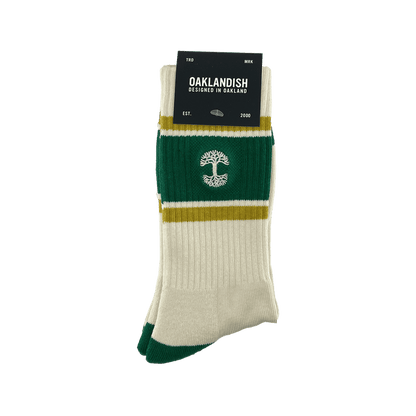 A brand new pair of Striped Classic Socks from Oaklandish, designed in a cream and green color palette with mustard yellow stripes, is neatly folded and adorned with a black cardboard tag. Made from 70% cotton, these crew socks feature a distinctive white tree emblem in the center. The tag reads "Oaklandish, Designed in Oakland," followed by "EST." and "2000." They are presented against a white background.