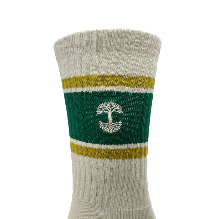 A close-up view of the Striped Classic Sock by Oaklandish, offered in adult sizes. This beige crew sock features a green band with yellow borders around the calf area, showcasing a white tree emblem knitted in the center. Made from 70% cotton, it has a ribbed texture and appears to be mid-calf length.