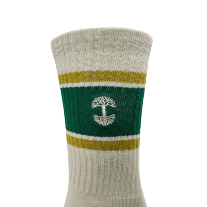 A close-up view of the Striped Classic Sock by Oaklandish, offered in adult sizes. This beige crew sock features a green band with yellow borders around the calf area, showcasing a white tree emblem knitted in the center. Made from 70% cotton, it has a ribbed texture and appears to be mid-calf length.