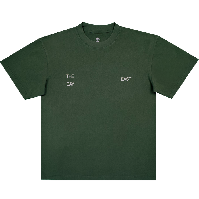 The East Bay Tee by Oaklandish is a dark green short-sleeve T-shirt from the Oaklandish x Toro y Moi collaboration, featuring small white text on the chest that reads "THE BAY" on the left and "EAST" on the right, separated by space. Inspired by Chaz Bear's unique style, this shirt is shown laid flat against a white background.