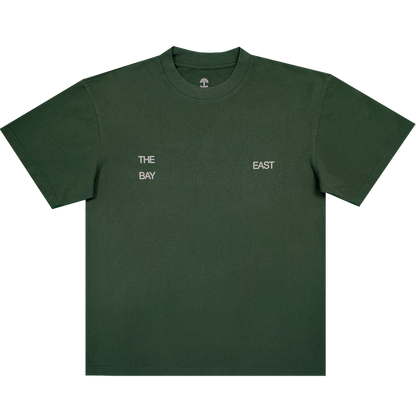 The East Bay Tee by Oaklandish is a dark green short-sleeve T-shirt from the Oaklandish x Toro y Moi collaboration, featuring small white text on the chest that reads "THE BAY" on the left and "EAST" on the right, separated by space. Inspired by Chaz Bear's unique style, this shirt is shown laid flat against a white background.