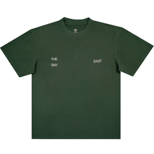 The East Bay Tee by Oaklandish is a dark green short-sleeve T-shirt from the Oaklandish x Toro y Moi collaboration, featuring small white text on the chest that reads "THE BAY" on the left and "EAST" on the right, separated by space. Inspired by Chaz Bear's unique style, this shirt is shown laid flat against a white background.