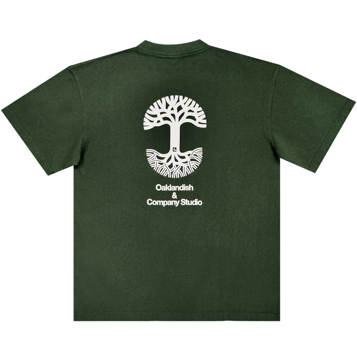 Certainly! Here’s the rewritten sentence using your product data: The Oaklandish East Bay Tee features a dark green color and a striking large white tree graphic with roots encircling a cityscape. Centered beneath the design on the back is the text "Oaklandish & Company Studio" in white. This exclusive Chaz Bear t-shirt is part of a special Toro y Moi collaboration with Oaklandish.