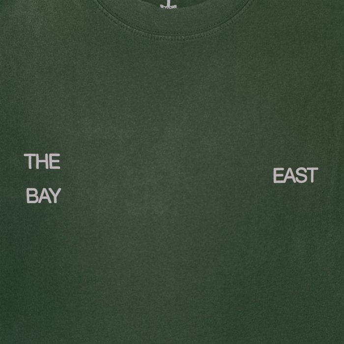 The East Bay Tee by Oaklandish is a dark green t-shirt featuring white, spaced-out text: "THE" above "BAY" on the left chest and "EAST" on the right chest. This design subtly nods to a Toro y Moi collaboration and Chaz Bear’s classic t-shirt style.