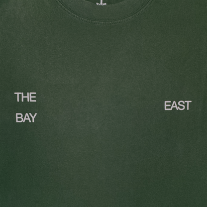 The East Bay Tee by Oaklandish is a dark green t-shirt featuring white, spaced-out text: "THE" above "BAY" on the left chest and "EAST" on the right chest. This design subtly nods to a Toro y Moi collaboration and Chaz Bear’s classic t-shirt style.