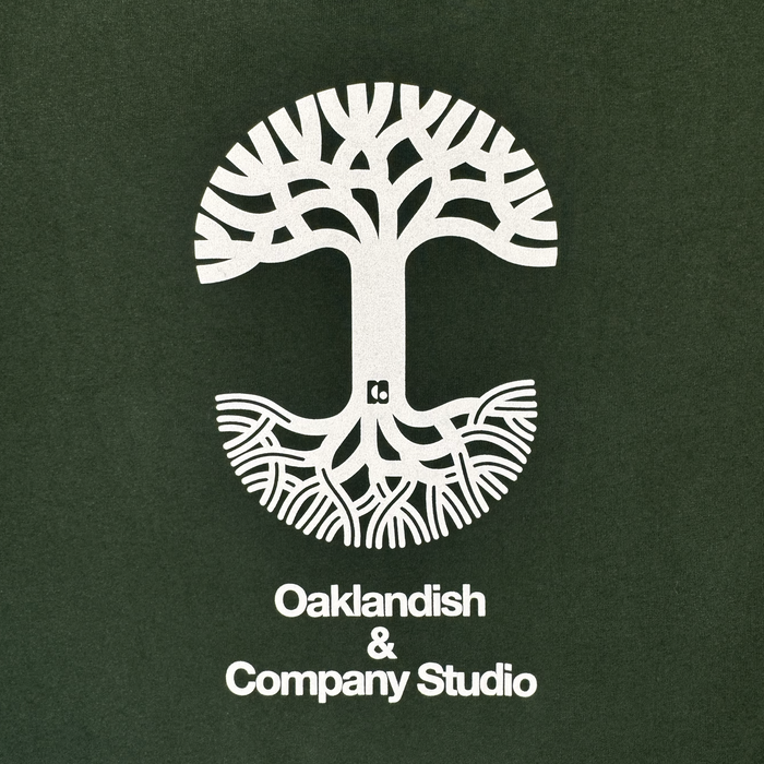 A white tree with stylized branches and roots creates a circular motif against a dark green background, echoing Oaklandish’s iconic design aesthetic. Beneath the tree, crisp white lettering spells out: “Oaklandish & Company Studio.” This clean, modern style serves as the perfect backdrop for The East Bay Tee—a limited-edition release from Oaklandish in collaboration with Toro y Moi’s Chaz Bear.