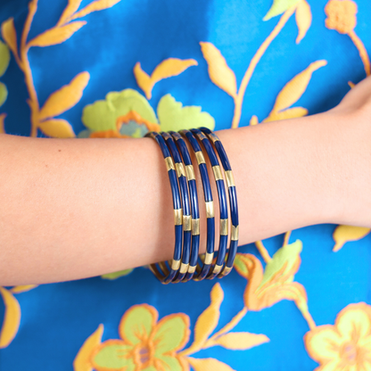 A stack of multiple bangle bracelets with dark blue and gold striped patterns, worn on a person's wrist against a bright blue floral fabric background.