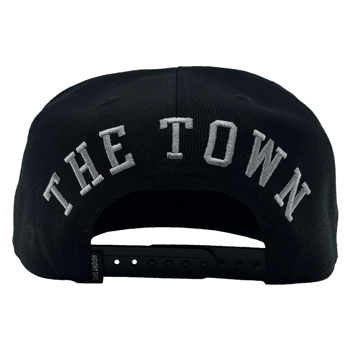 The rear view of "The Town Aft Snapback" by Oaklandish features "THE TOWN" prominently embroidered in large white letters. The hat's adjustable snapback closure, a small label tag on the side, and an embroidered Oaklandish tree logo are also visible.