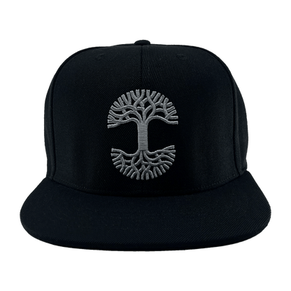 The Town Aft Snapback by Oaklandish is a black hat featuring a wide, flat brim. The front prominently displays a white embroidered Oaklandish tree logo with symmetrical branches and roots, reminiscent of the tree of life symbol. The background is solid green and includes an adjustable snapback for the perfect fit.