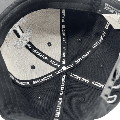 The image shows the inside of "The Town Aft Snapback," a black hat from Oaklandish, with the brand name "OAKLANDISH" repeatedly printed on the interior seam taping. The under-visor features a textured and patterned design in light gray, including a partial embroidered Oaklandish tree logo. The overall material appears to be fabric.