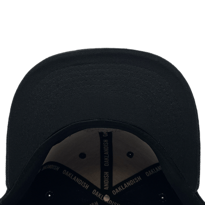 The Town Aft Snapback by Oaklandish, viewed from a top-down perspective, reveals the underside of the brim with detailed stitching. Inside, a black band with "OAKLANDISH" printed in white is sewn around the interior. This adjustable snapback also features an embroidered Oaklandish tree logo. The background is plain white.