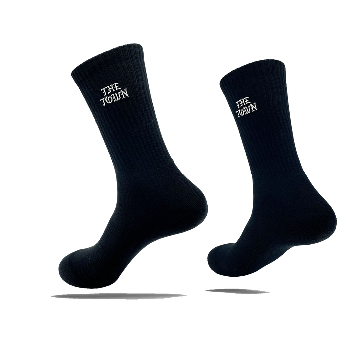 Image of two black crew socks, each showcasing the white text "THE TOWEL" near the top. These stylish crew socks, named The Town Old English Sock by Oaklandish, are one size fits most and appear to be floating against a transparent background, casting a shadow beneath each sock. The material features a ribbed texture at the top.
