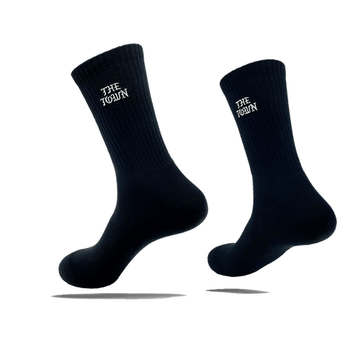 Image of two black crew socks, each showcasing the white text "THE TOWEL" near the top. These stylish crew socks, named The Town Old English Sock by Oaklandish, are one size fits most and appear to be floating against a transparent background, casting a shadow beneath each sock. The material features a ribbed texture at the top.