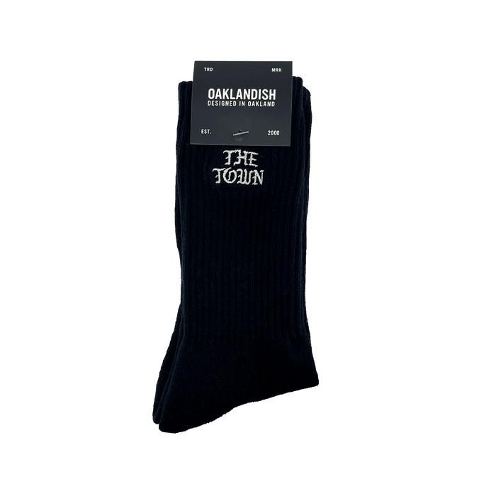 The Town Old English Sock, designed by Oaklandish, is a pair of black crew socks featuring "THE TOWN" embroidered in white lettering near the top. The packaging includes a black label with "OAKLANDISH DESIGNED IN OAKLAND," the Nike logo, and additional text stating "FOR MEN," "ONE SIZE FITS MOST," and "EST. 2000.