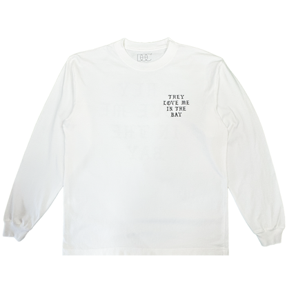 A heavyweight cotton long sleeve t-shirt by By DOC, the They Love Me LS Tee By Doc, is shown in men’s sizing laid flat against a plain white background. The only visible design is small black text on the left chest that reads, “THEY LOVE ME IN THE BAY.” There are no other embellishments or graphics.