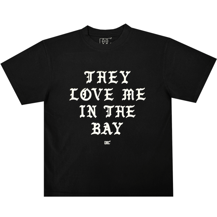 They Love Me Tee By Doc from By DOC, shown laid flat on a plain white background. This classic-fit black t-shirt is crafted from heavyweight cotton and features bold, white Old English-style lettering across the front that reads, “THEY LOVE ME IN THE BAY.” A small white By DOC logo sits just below the text at the center.