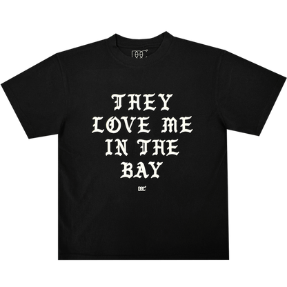 They Love Me Tee By Doc from By DOC, shown laid flat on a plain white background. This classic-fit black t-shirt is crafted from heavyweight cotton and features bold, white Old English-style lettering across the front that reads, “THEY LOVE ME IN THE BAY.” A small white By DOC logo sits just below the text at the center.