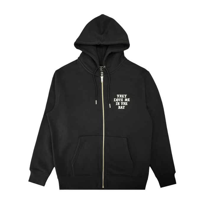 A black They Love Me Zip By Doc hoodie from By DOC, made with heavyweight fleece and designed with front pockets and a hood. The left chest features white, all-caps text that reads “THEY LOVE ME IN THE BAY.” The hoodie is displayed flat on a white background.