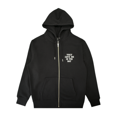 A black They Love Me Zip By Doc hoodie from By DOC, made with heavyweight fleece and designed with front pockets and a hood. The left chest features white, all-caps text that reads “THEY LOVE ME IN THE BAY.” The hoodie is displayed flat on a white background.