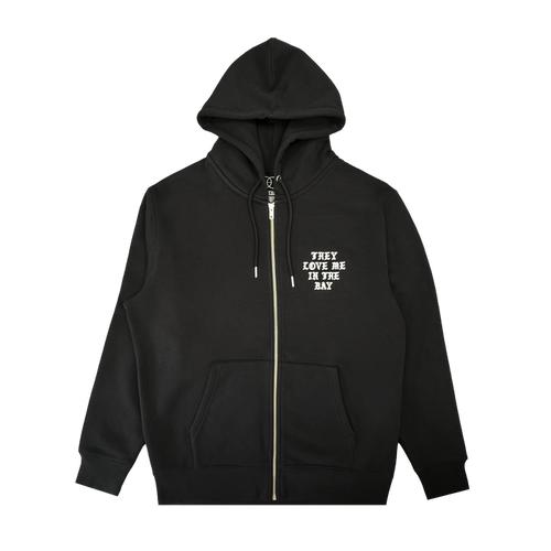 A black They Love Me Zip By Doc hoodie from By DOC, made with heavyweight fleece and designed with front pockets and a hood. The left chest features white, all-caps text that reads “THEY LOVE ME IN THE BAY.” The hoodie is displayed flat on a white background.