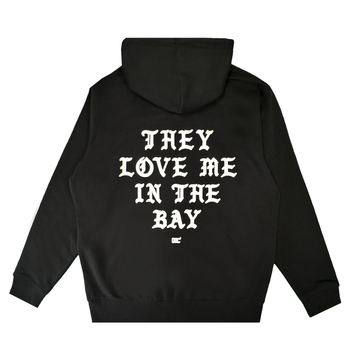 A black heavyweight fleece zip hoodie from By DOC’s They Love Me Zip collection, featuring white gothic-style text on the back that reads “THEY LOVE ME IN THE BAY” with a small white logo beneath it, laid flat on a white background.