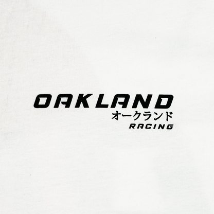 The Tuner LS Tee from Oaklandish is a white, 100% cotton long sleeve shirt with bold black "OAKLAND" in caps and Japanese katakana "オークランド." Below, "RACING" appears in smaller letters, slightly tilted right. It offers a classic fit with a modern and simple design.