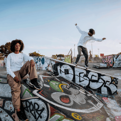 Sporting an Oaklandish Tuner LS Tee, a person sits on a graffiti-covered ramp at an open skate park, watching another skateboarder in white and brown navigate a slope under the clear sky.