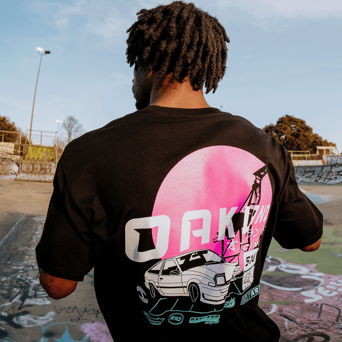 In a graffiti-filled skate park, a person with curly hair wears the black Oaklandish Tuner Tee, featuring 100% cotton fabric with a retro car beneath a pink sun and bold "OAKLAND," surrounded by trees and lampposts in the design.