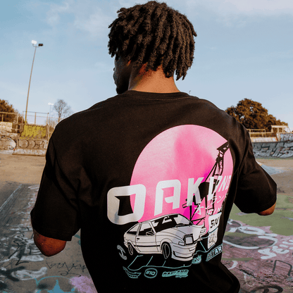In a graffiti-filled skate park, a person with curly hair wears the black Oaklandish Tuner Tee, featuring 100% cotton fabric with a retro car beneath a pink sun and bold "OAKLAND," surrounded by trees and lampposts in the design.