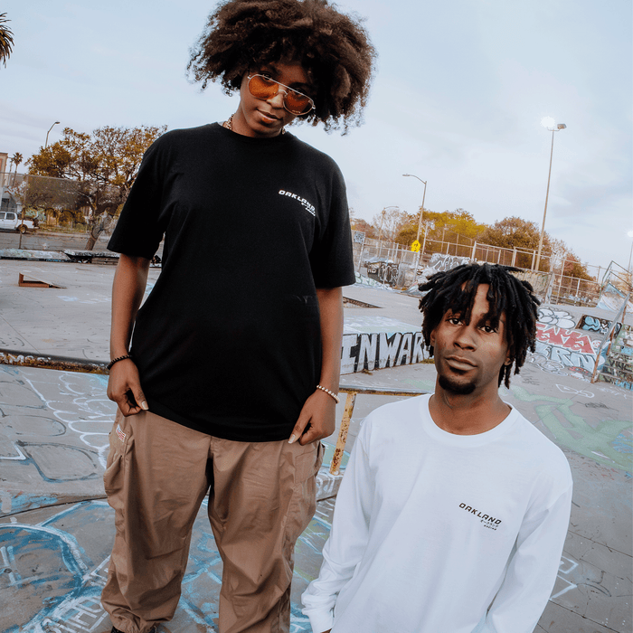 Two graffiti park visitors flaunt Oaklandish style: one in a black Tuner Tee and beige pants with sunglasses, the other near a ramp in a white long-sleeve tee. Both 100% cotton tees feature the same logo. Trees and a cloudy sky provide the backdrop.
