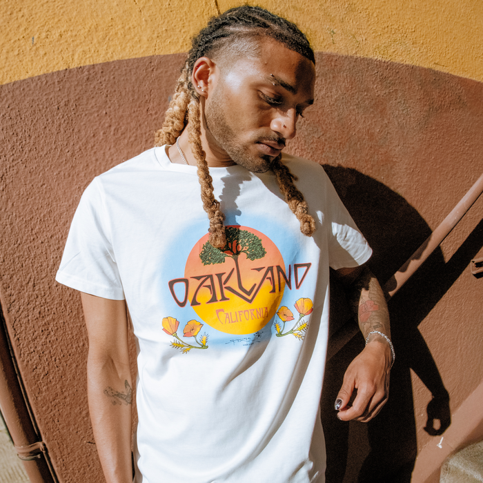 Wearing the Oaklandish Vanguard Tee, featuring "Oakland California" with a sunset, trees, and flowers graphic, a person with long blond-tipped braids stands against a yellow and brown wall, looking down with one hand by their side.
