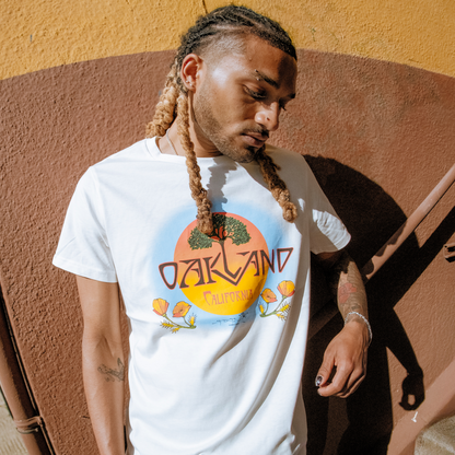 Wearing the Oaklandish Vanguard Tee, featuring "Oakland California" with a sunset, trees, and flowers graphic, a person with long blond-tipped braids stands against a yellow and brown wall, looking down with one hand by their side.