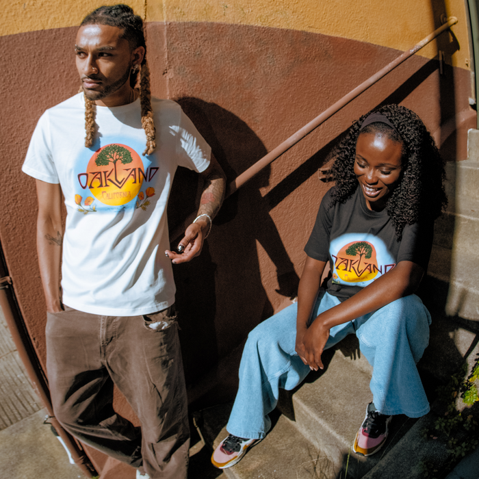 A man stands and a woman sits on steps beside him, both wearing the Oaklandish Vanguard Tee, which features bold album artwork-inspired graphics with colorful sunrise motifs. Sunlight casts strong shadows on the textured wall behind them.