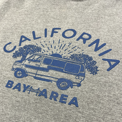The Wavy Nostalgia Hoodie by Never Elsewhere features a Gunmetal Heather gray pullover design with a blue graphic depicting a van beneath two large trees. A sun with rays shines behind the van, and the words "CALIFORNIA" and "BAY AREA" surround the graphic, with a bridge illustration located below the "BAY AREA" text. Available in men's sizing.